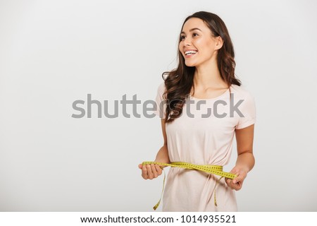 Portrait of a happy young asian woman holding measuring tape around her waist and looking away at copy space over white background Royalty-Free Stock Photo #1014935521