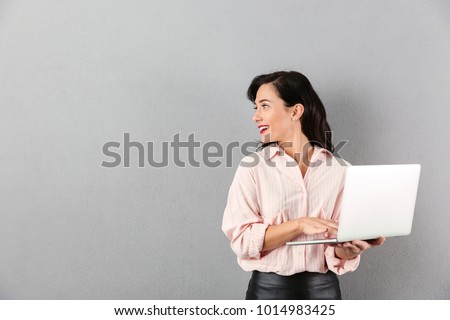 Portrait of a smiling businesswoman using laptop computer while standing and looking away at copy space isolated over gray background