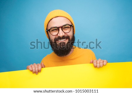 Smiling bearded man in hat and glasses holding yellow banner on yellow background. 