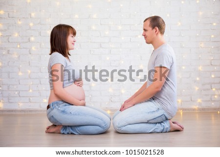family, parenthood and happiness concept - portrait of pregnant couple sitting on the floor and looking each other over white brick wall with christmas lights