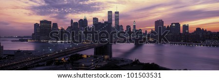 This is the Brooklyn Bridge over the East River and the Manhattan skyline at sunset. There is a purple cast to the sky and the lights to the buildings are starting to come on.