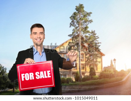 Real estate agent holding sign with text FOR SALE and key in front of country house outdoors