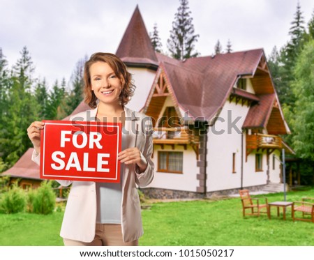 Real estate agent holding sign with text FOR SALE in front of country house outdoors