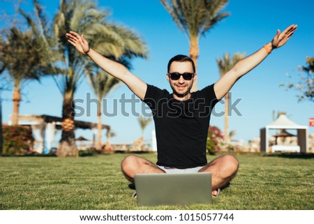 Handsome man sitting on the grass and use laptop with victory raised hands on palm trees background. Summer vocation.