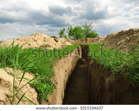 Inside the trench, excavated with help of special equipment on the military training ground (firing field), green grass and white sand on the sides, green trees, gloomy sky