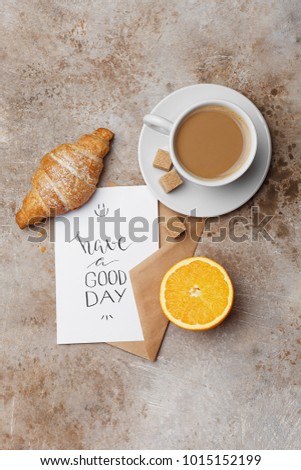 Morning cup of coffee, croissant, fresh orange and card "have a good day" over beige concrete background. Healthy delicious breakfast. Overhead view, flat lay