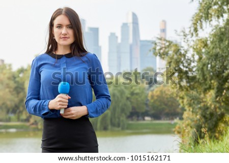 Young woman reporter stands with microphone against pond, green trees and skyscrapers.