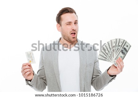 Picture of handsome young businessman standing isolated over white background. Looking camera holding credit card and money while winking.