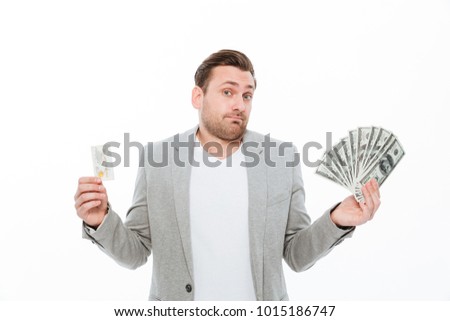 Picture of confused young businessman standing isolated over white background. Looking camera choosing between credit card and money.