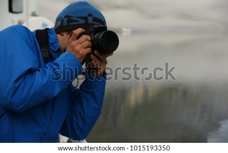 Young man in blue jacket  with photo camera in hands making photo, on background of fog over water of Norwegian fjord, closeup 