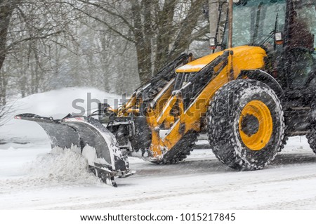 Yellow tractor with snowplow removing snow during heavy snowfall. Winter time street maintenance in hard weather conditions. Blizzard, traffic