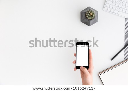 Hand holding blank smartphone with blank screen over office desk with supplies and a cactus.