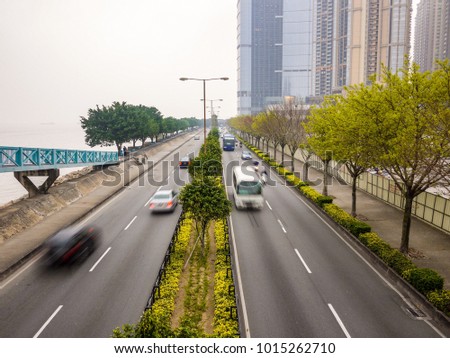 Median divider with green trees and bush plants in the middle of urban road. Cars and bus driving on highway by the sea with pedestrian sidewalk on side modern skyscrapers in distance. Macau, China. Royalty-Free Stock Photo #1015262710