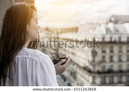 Young attractive brunette enjoying a cup of coffee in the morning on a balcony in France with beautiful Paris city architecture in the background