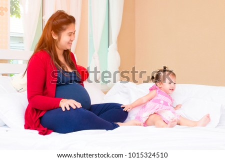 The beautiful pregnant mother with red knitting sweater playing with cute little daughter in pink dress are smiling while spending time together in the bedroom of the home in the morning. Happy family