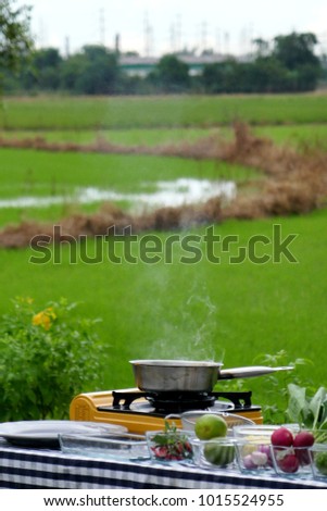 Blurry photo of cooking at rice field.  Cooking smoke emitting from cooking pan on yellow picnic stove/portable gas stove.