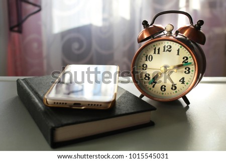 Alarm clock and mobile smart phone on desk near the window with morning light and copy space, waiting for an important call, count down, vintage tone. timing management and reading book concept.
