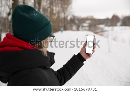A girl is looking at the phone in the background of a park and a winter village.Holds the phone with your left hand.