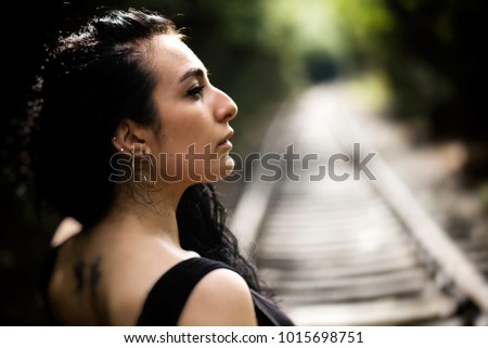 Portrait in profile of an attractive lonely young woman  with long black hair, wearing black dress on background the railway tracks, summer nature outdoor, tone colour
