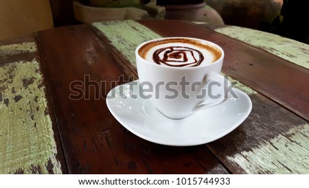 A cup of coffee latte with spiral shape of chocolate sauce on top with white cup and saucer on the vintage wooden brown and green stripe pattern round table with ancient pottery on the background