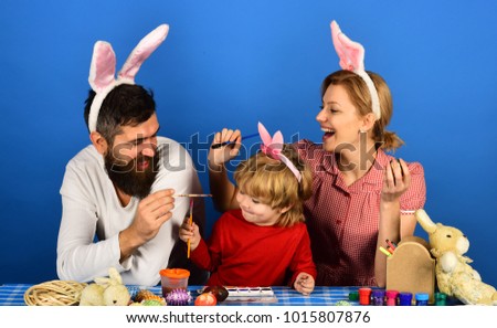 Family members painting eggs on blue background. Holiday spirit and joy concept. Man, woman and kid wearing bunny ears. Mother, father and daughter with smiling and curious faces preparing for Easter.