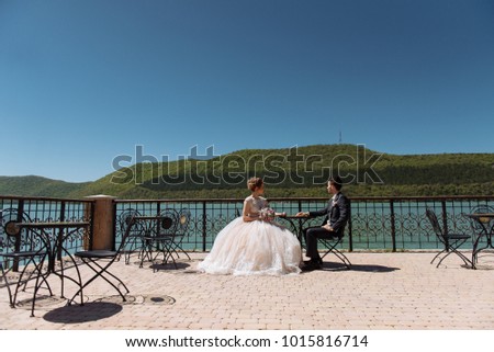 The bride and groom are sitting at a table in front of a lake and mountains and holding hands