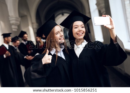 Two girls in black mantles are making selfie. They are university graduates. They are happy about this. They are in a good mood.