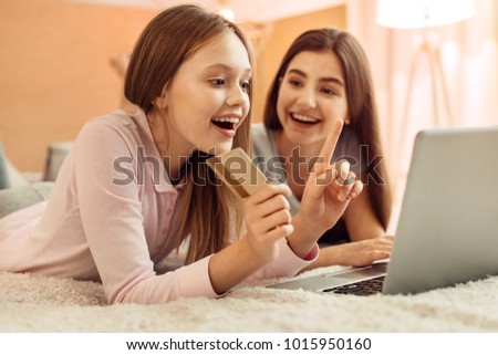 Incurable shopaholic. Excited pretty teenage girl lying on the bed next to her elder sister and choosing an item in an online shop while holding a golden credit card Royalty-Free Stock Photo #1015950160