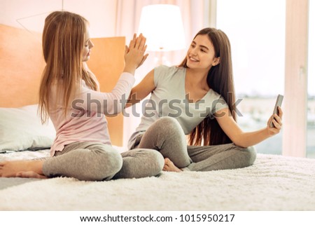 Great job. Adorable teenage sisters sitting on the bed in their room and giving each other a high-five after taking good selfies together