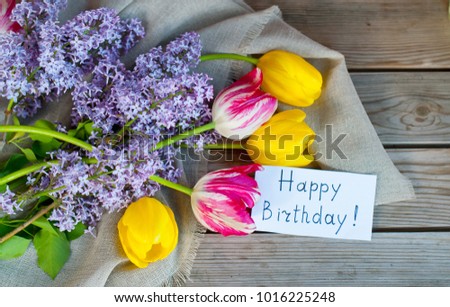 A bouquet of tulips and lilacs with an inscription happy birthday on a wooden background. Festive postcard. Decorative, spring concept.