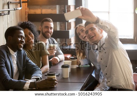 Diverse friends making selfie on smartphone together in cafe, young man holding phone shooting video or taking photo on mobile of african and caucasian millennial people sitting at coffee shop table Royalty-Free Stock Photo #1016243905