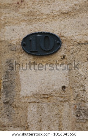 Close up view of a small oval metal plate fixed on an exterior wall house. Number 10 raised written. Black plate fixed on a beige stone facade. Abstract view of the number ten. Simple figure.    