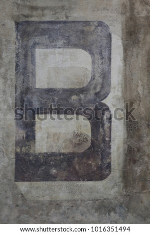 Capital letter B written on a grey ancient wall. Dark painted character on a flat vertical surface. Simple abstract picture. Symbol of the alphabet. Brown, black, grey and beige colors.  