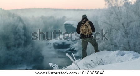 A brutal tourist man walks through a snow-covered forest against the background of a river and rocks at dawn