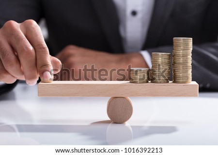 Close-up Of A Businessperson's Hand Balancing Stacked Coins On Wooden Seesaw With Finger Over Desk