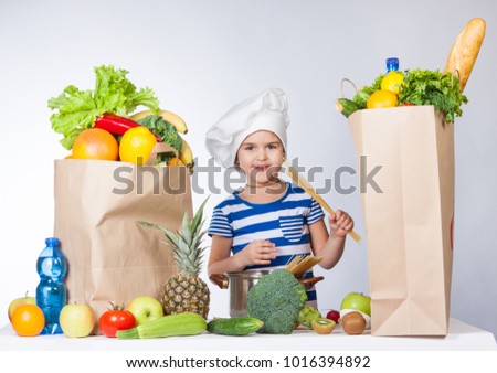 Happy little girl in the hat of the chef with big bags of food. A variety of fresh fruits and vegetables in bags on the table. Health food. The child holds the spaghetti