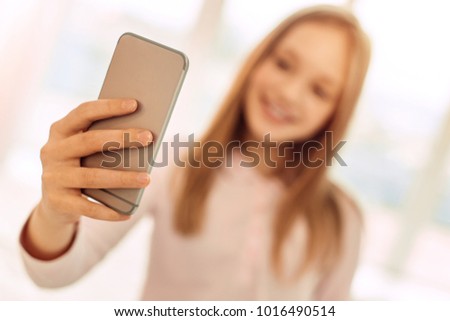 Helpful gadget. The focus being on the phone in the hands of a pretty fair-haired teenage girl standing in the room and taking a selfie while smiling