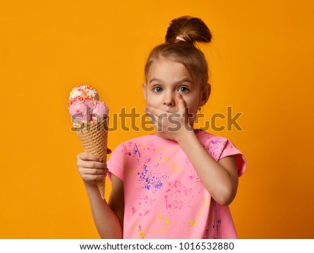 Pretty baby girl kid hold big banana and strawberry ice cream in waffles cone and close mouth with hand on yellow background 