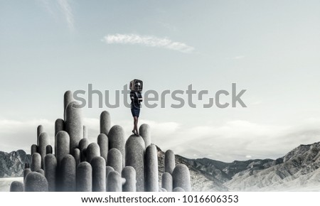 Business woman in suit with an old TV instead of head keeping arms crossed while standing on the top of stone columns with beautiful landscape on background.