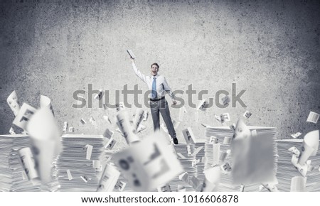 Businessman keeping hand with book up while standing among flying documents with grey background. Mixed media.