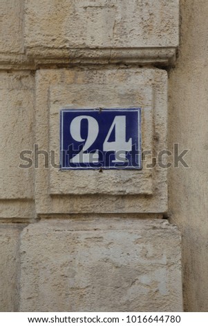 Close uo view of the number 24 written in white on a blue metallic rectangle. Element fixed on an house beige stone wall in a street in France. Graphic urban picture. Simple sign. Geometric design. 