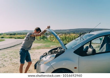 Young man looking under the hood of breakdown car outdoors. Transportation and vehicle concept Royalty-Free Stock Photo #1016692960