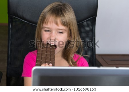 Little girl sitting at the computer and eating chocolate.