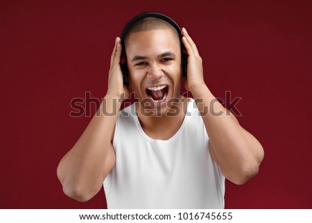 Picture of charismatic emotional mixed race guy having fun, listening to music in black headphones, expressing positivity and screaming while singing along with favourite tracks, feeling joyful