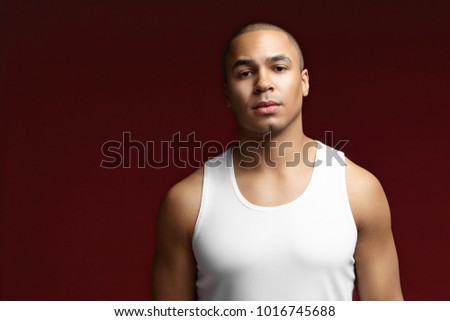 People, style, lifestyle and fashion concept. Picture of good looking stylish mixed race sportsman wearing white sleeveless shirt posing in studio, having serious confident facial expression