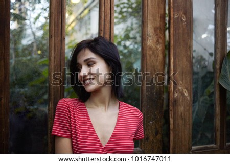 Picture of fashionable young brunette European lady wearing stylish red dress with white stripes posing outdoors at wooden door with mirrored surface, looking away with positive happy smile