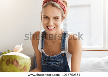 Pretty female dressed in fashionable clothes, has pleasant smile on face, being satisfied with something, drinks coconut cocktail. Positive woman spends free time at cafeteria in tropical country
