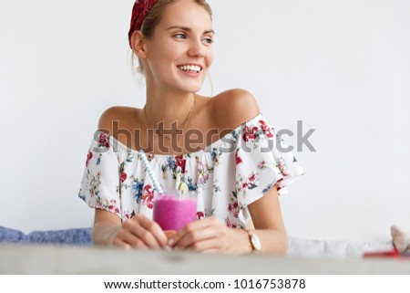 Cropped shot of pretty happy woman drinks smoothie in cafeteria, enjoys spare time, looks thoughtfully aside, has happy positive expression. Smiling young female with beverage at cozy cafeteria