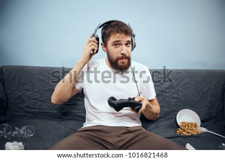  man in headphones in hand joystick sitting on sofa with garbage on blue background                              