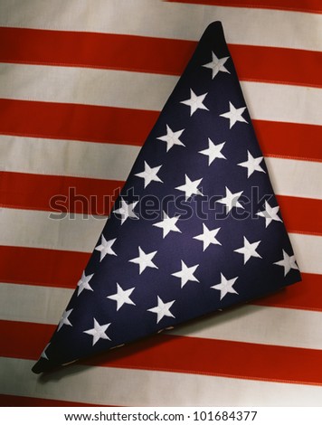 This shows a triangularly folded American flag sitting on top of another American flag which only shows its red and white stripes going in a horizontal direction.